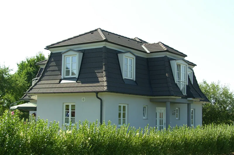 Weißes Haus mit dunklem Mansarddach und Gauben hinter einer Hecke